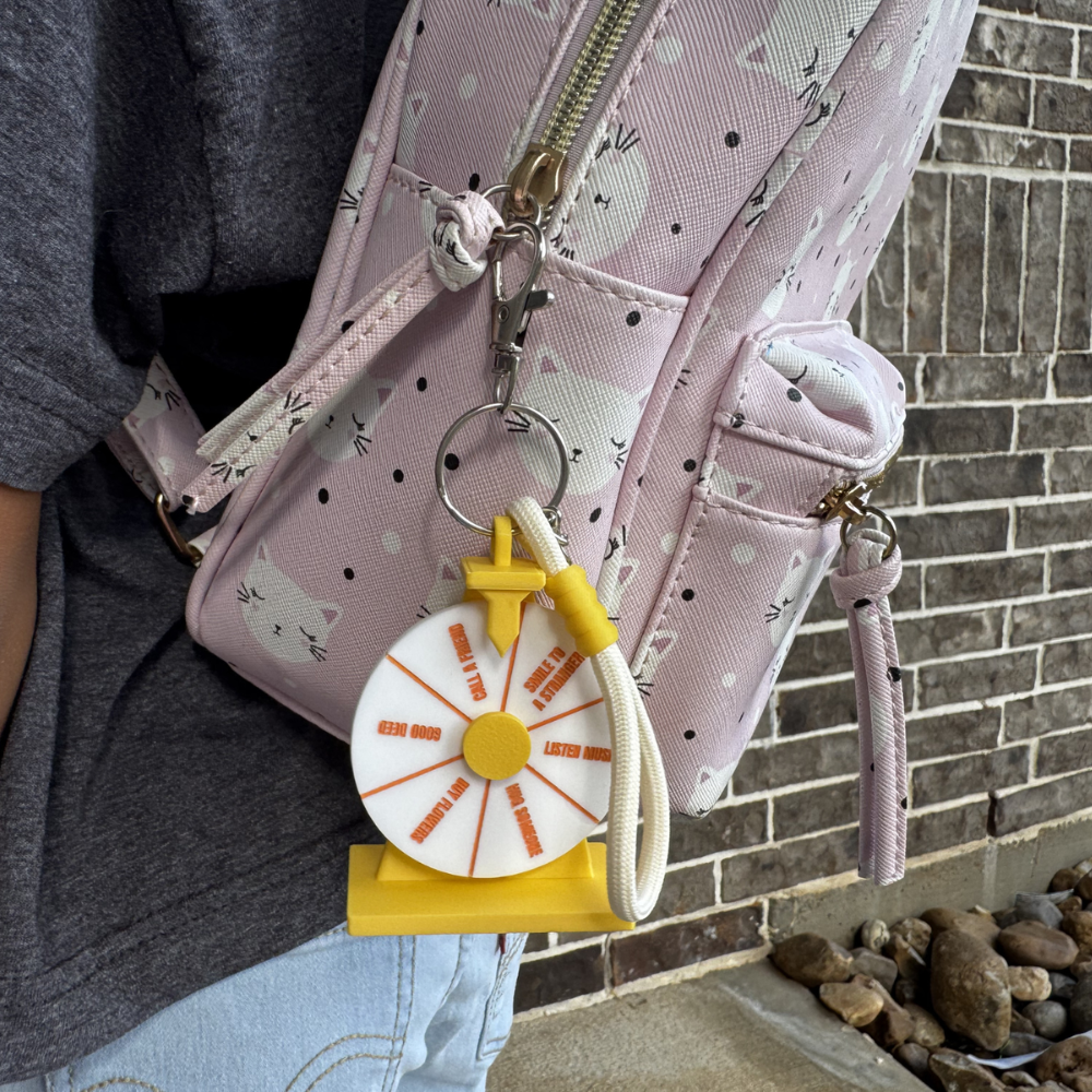 A pink patterned crossbody bag with a yellow and white spinner keychain charm, shown worn on a person against a brick wall background.