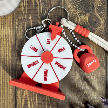 A fitness-themed keychain set featuring a red and white wheel labeled with recovery types like rest, lift, walk, milk, protein, and hydrate, attached to a silver key ring, red weight ball, and white rope on a rustic wooden background.